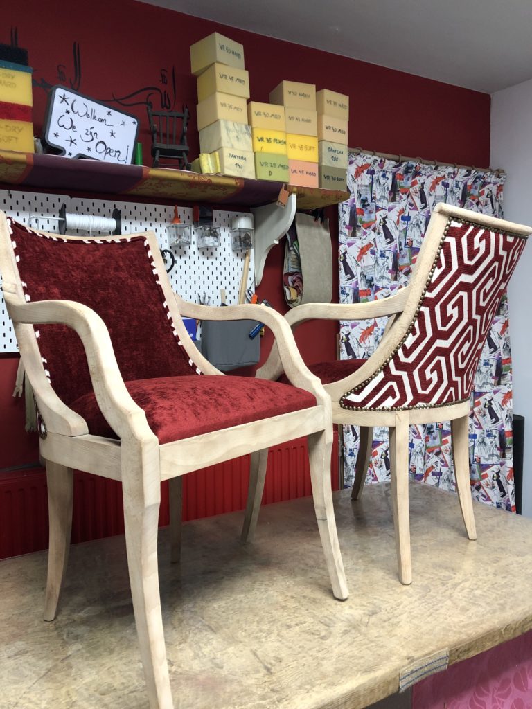 twee vintage stoelen staan op een werktafel. Ze zijn opnieuw geherstoffeerd in een roodkleurige stof. De achterzijde van de stoelen is in een andere stof bekleed.